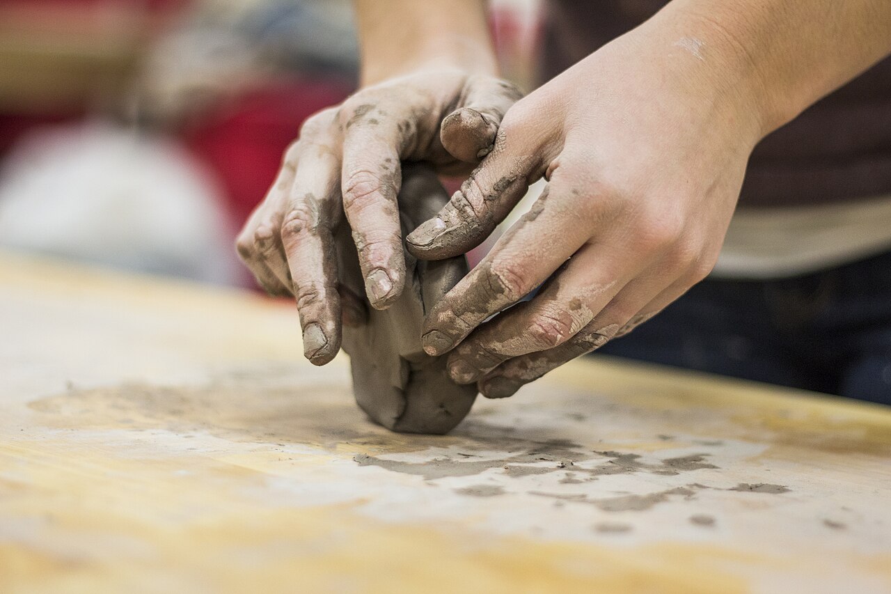 Two clay-covered hands shaping a pot on a workbench, used as reference craft photography